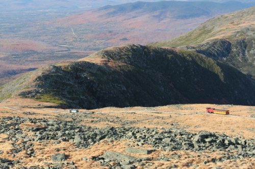 A mountainous landscape with rocky terrain and a winding road, featuring two small vehicles in the distance.