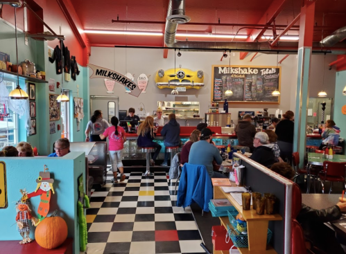 A colorful diner interior with checkered floors, patrons at tables, and a counter serving milkshakes.