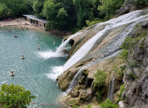 A scenic view of waterfalls cascading into a turquoise pool, surrounded by lush greenery and a sandy beach area.