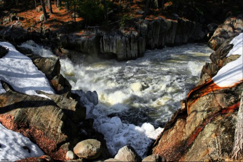 A rushing river flows through rocky terrain, surrounded by snow and trees in a natural landscape.