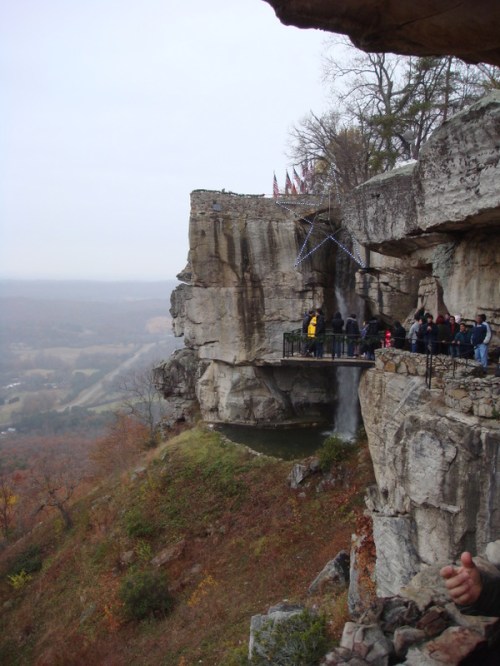 A rocky cliff with a viewing platform, surrounded by autumn foliage and a distant valley under a cloudy sky.