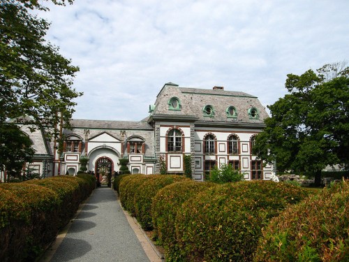 A grand, historic building with ornate architecture, surrounded by neatly trimmed hedges and a cloudy sky.