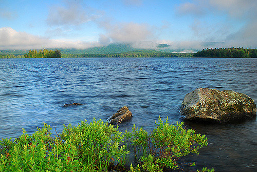 A serene lake scene with gentle waves, rocky shore, and misty mountains in the background under a clear blue sky.