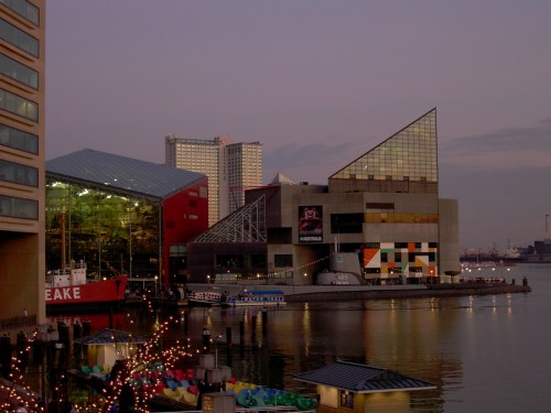 A waterfront view of modern buildings and boats at dusk, reflecting in calm water with city lights in the background.