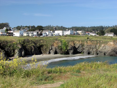 Coastal view of a small town with white buildings, cliffs, and a calm blue water inlet surrounded by greenery.
