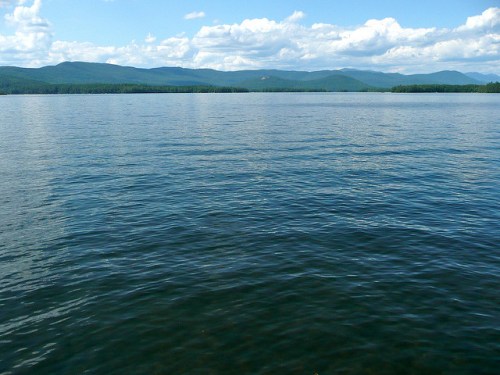 A serene lake with calm waters, surrounded by mountains and a blue sky with fluffy clouds.