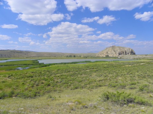 A scenic landscape featuring a rocky outcrop, green fields, and a calm lake under a blue sky with fluffy clouds.