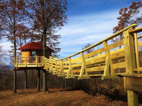 A wooden treehouse with a red roof, connected by a curved wooden walkway, surrounded by autumn trees and blue sky.