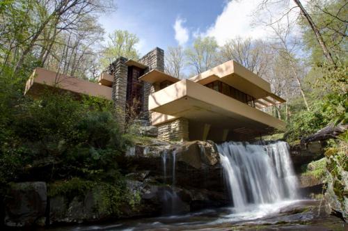 A modern house with cantilevered roofs, nestled among trees beside a waterfall and rocky landscape.