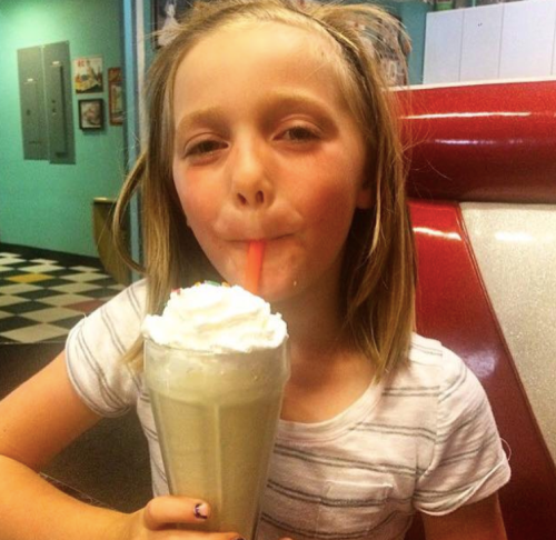 A young girl with blonde hair enjoys a milkshake topped with whipped cream, sipping through a straw.
