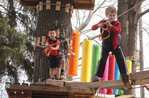 Three children navigate a ropes course, with colorful hanging tubes in the background and trees surrounding them.