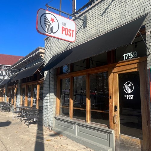 Exterior of "The Post" with a sign, large windows, and outdoor seating on a sunny day.