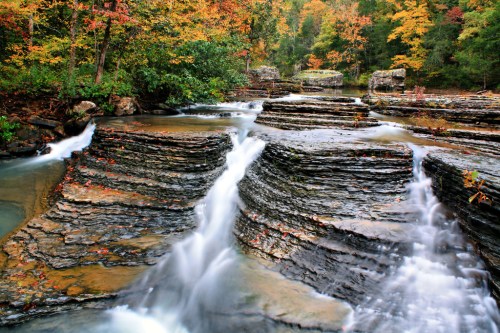 A serene waterfall cascades over layered rocks, surrounded by vibrant autumn foliage.