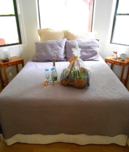 A neatly made bed with purple pillows, a gift basket, and two bottles on the side tables, in a bright room.