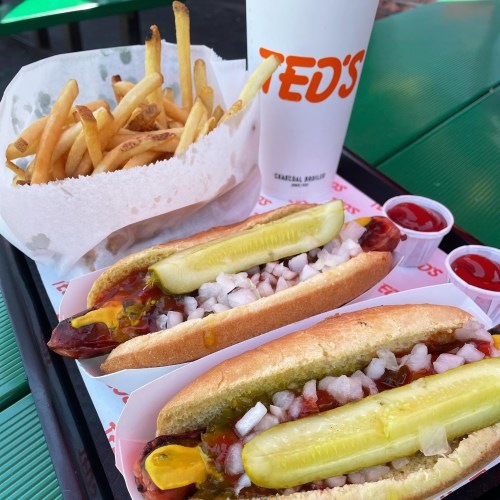 A tray with two hot dogs topped with pickles, onions, and condiments, alongside a serving of fries and a drink.