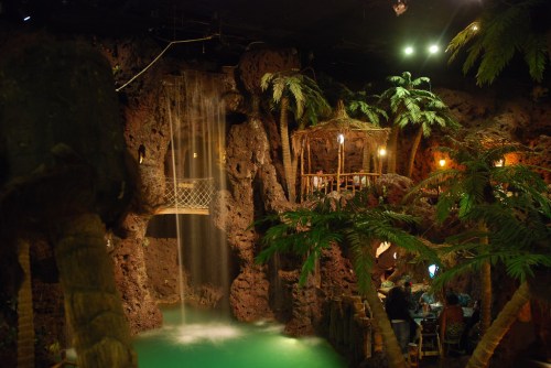 A tropical-themed indoor setting featuring a waterfall, lush palm trees, and a cozy seating area.