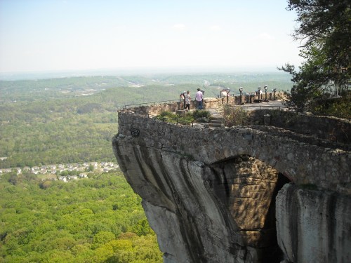 A scenic overlook with people enjoying the view from a rocky cliff, surrounded by lush green hills and a clear sky.