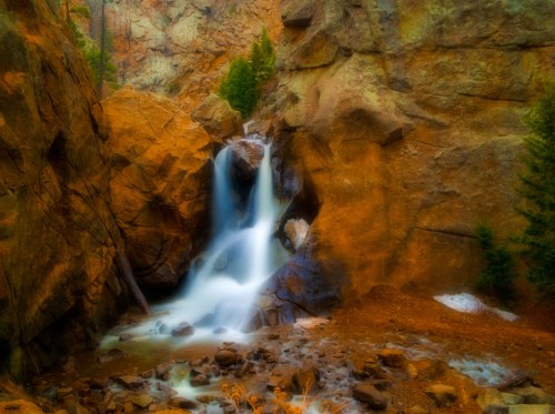 A serene waterfall cascades over rocks, surrounded by rugged cliffs and evergreen trees in a tranquil natural setting.