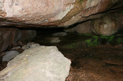 A dimly lit cave interior with large rocks, wet ground, and patches of green moss.