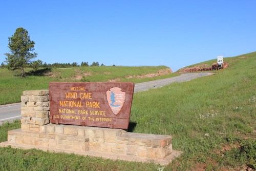 Welcome sign for Wind Cave National Park, with a road and green hills in the background under a clear blue sky.