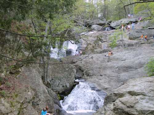 A scenic waterfall cascades over rocky terrain, with people enjoying the natural surroundings in a lush forest.