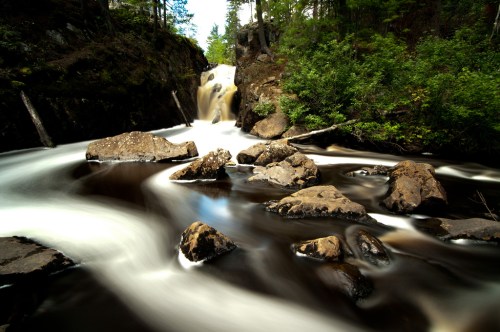 A serene river flows over rocks, surrounded by lush greenery and a waterfall in the background.