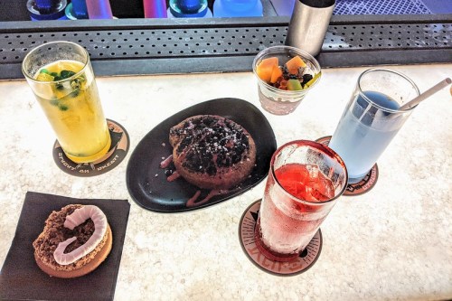 A table with five colorful drinks and a dessert, including a chocolate cake and a fruit cup, on coasters.