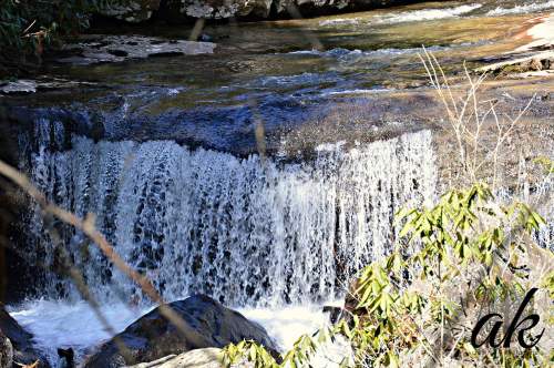 A small waterfall cascades over rocks, surrounded by greenery and flowing water in a serene natural setting.