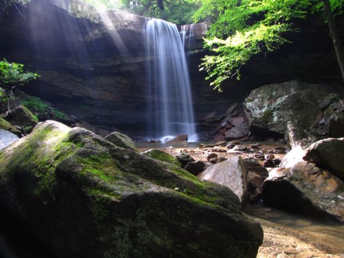 A serene waterfall cascades over rocks, surrounded by lush greenery and sunlight filtering through the trees.