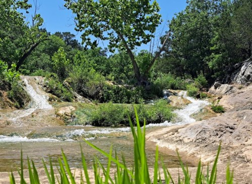 A serene landscape featuring a flowing stream, lush greenery, and rocky terrain under a clear blue sky.