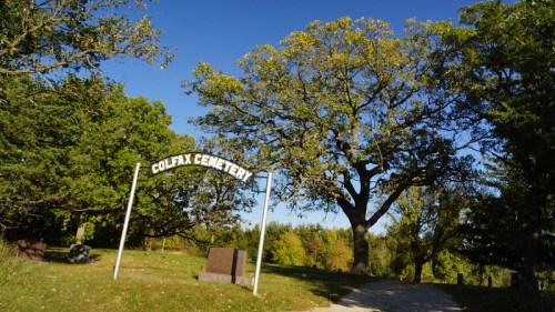 Sign for Colfax Cemetery surrounded by trees and a clear blue sky. Gravel path leads into the cemetery area.