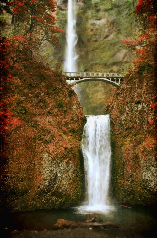 A scenic waterfall cascades between rocky cliffs, with a bridge arching above, surrounded by vibrant autumn foliage.