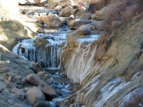 A frozen waterfall cascades over rocky terrain, surrounded by icy formations and smooth boulders.