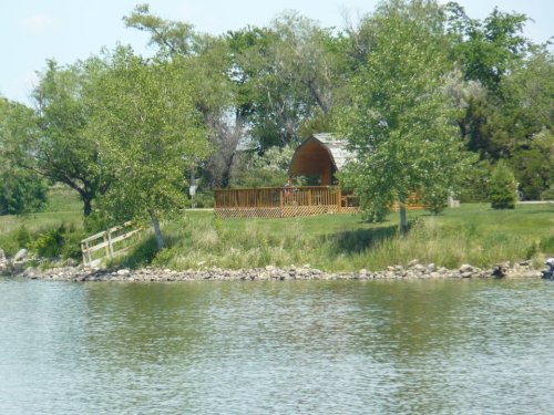 A serene lakeside scene featuring a wooden gazebo surrounded by green trees and grassy areas.