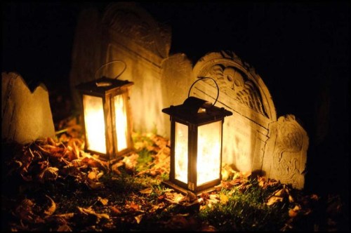 Two glowing lanterns beside old gravestones, surrounded by fallen leaves in a dimly lit setting.