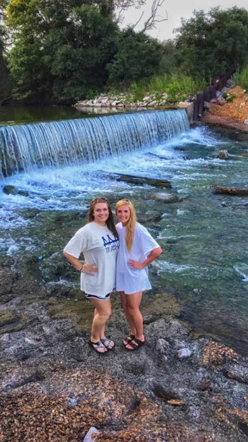 Two young women stand by a waterfall, smiling, with water flowing and greenery in the background.