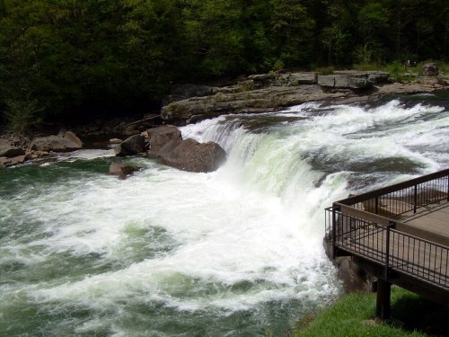 A waterfall cascades over rocks into a river, surrounded by lush greenery and a wooden viewing platform nearby.