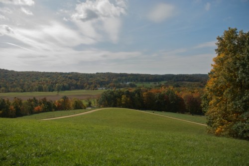 A scenic landscape featuring rolling hills, lush green grass, and autumn-colored trees under a partly cloudy sky.