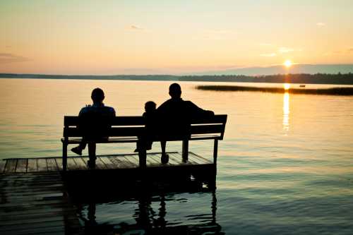 A family sits on a dock at sunset, overlooking a calm lake with reflections of the colorful sky.