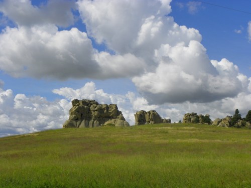 A grassy field with large rock formations under a blue sky filled with fluffy white clouds.