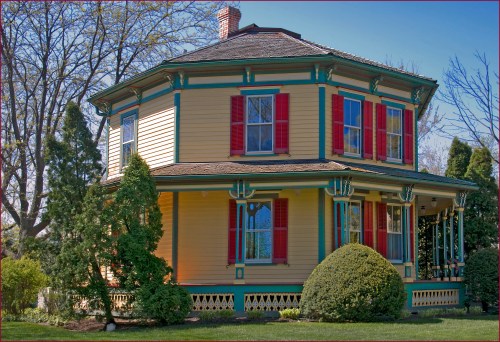 A colorful Victorian-style house with yellow siding, red shutters, and green trim, surrounded by lush greenery.