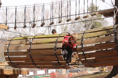 Two children play on a wooden climbing structure with ropes and logs in a park setting.