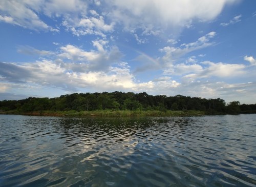 A serene lake scene with calm water reflecting clouds and a rainbow over a lush green island.