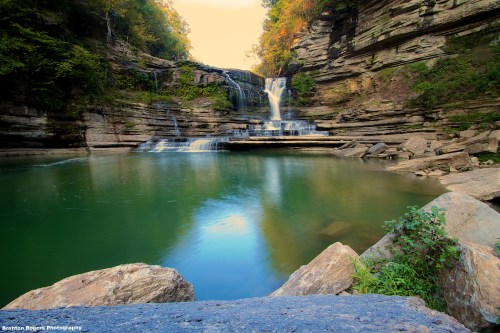 A serene waterfall cascades into a tranquil green pool, surrounded by rocky cliffs and autumn foliage.