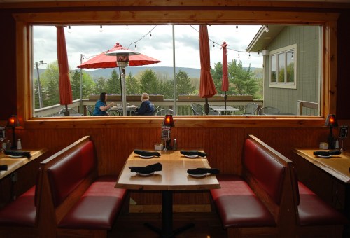 Cozy restaurant interior with red booths, overlooking a patio with umbrellas and a scenic view of trees and mountains.