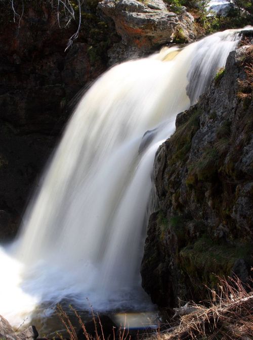 A cascading waterfall flows over rocky terrain, surrounded by lush greenery and natural textures.
