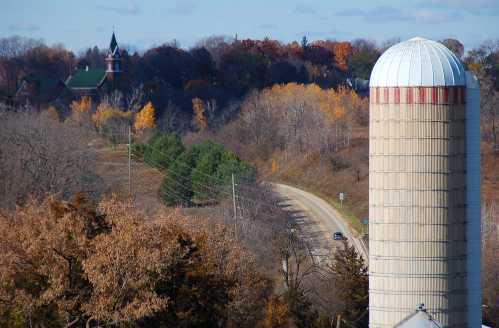 A rural landscape featuring a silo, winding road, and autumn trees with a church steeple in the background.