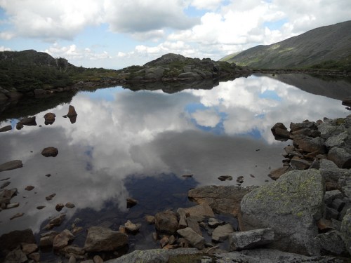 A serene landscape featuring a calm lake reflecting clouds and rocky terrain under a blue sky.