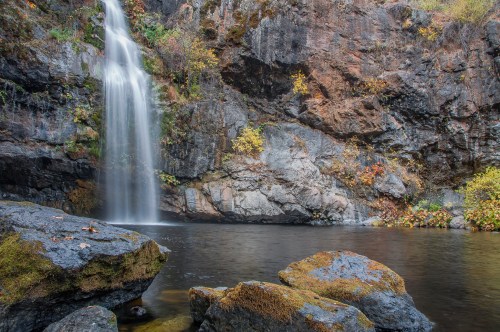 A serene waterfall cascades over rocky cliffs into a calm pool, surrounded by autumn foliage and textured stones.