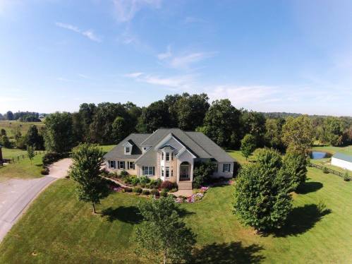 Aerial view of a large house surrounded by greenery and trees, with a driveway and a pond in the background.